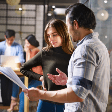 Male in mid thirties is explaining and gesturing towards a paper held out in front of him while a female in mid thirties looks at the paper with approval