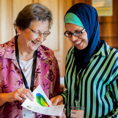 Elderly white woman holds a brightly colored poster for an elderly BIPOC woman with headscarf to reacd